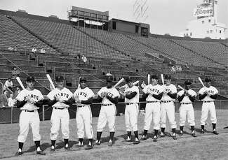 The original San Francisco Giants lineup stands in line in white uniforms and holds bats in their empty stadium.  In the background above the stand you can see the sign of the Hamm brewery. 