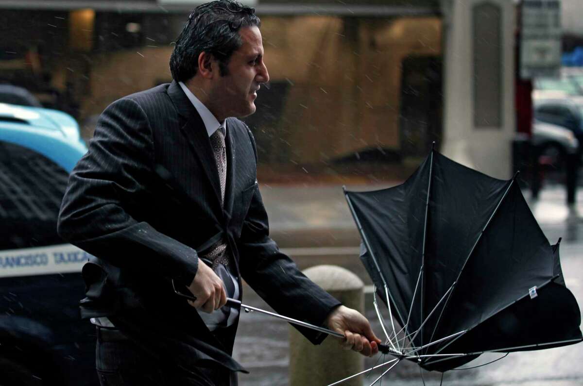 一名男子走在土耳其街上，他的雨伞在强风吹过旧金山市中心后倒塌了。另一场由大气河流驱动的风暴将抵达加州，带来另一轮强风。