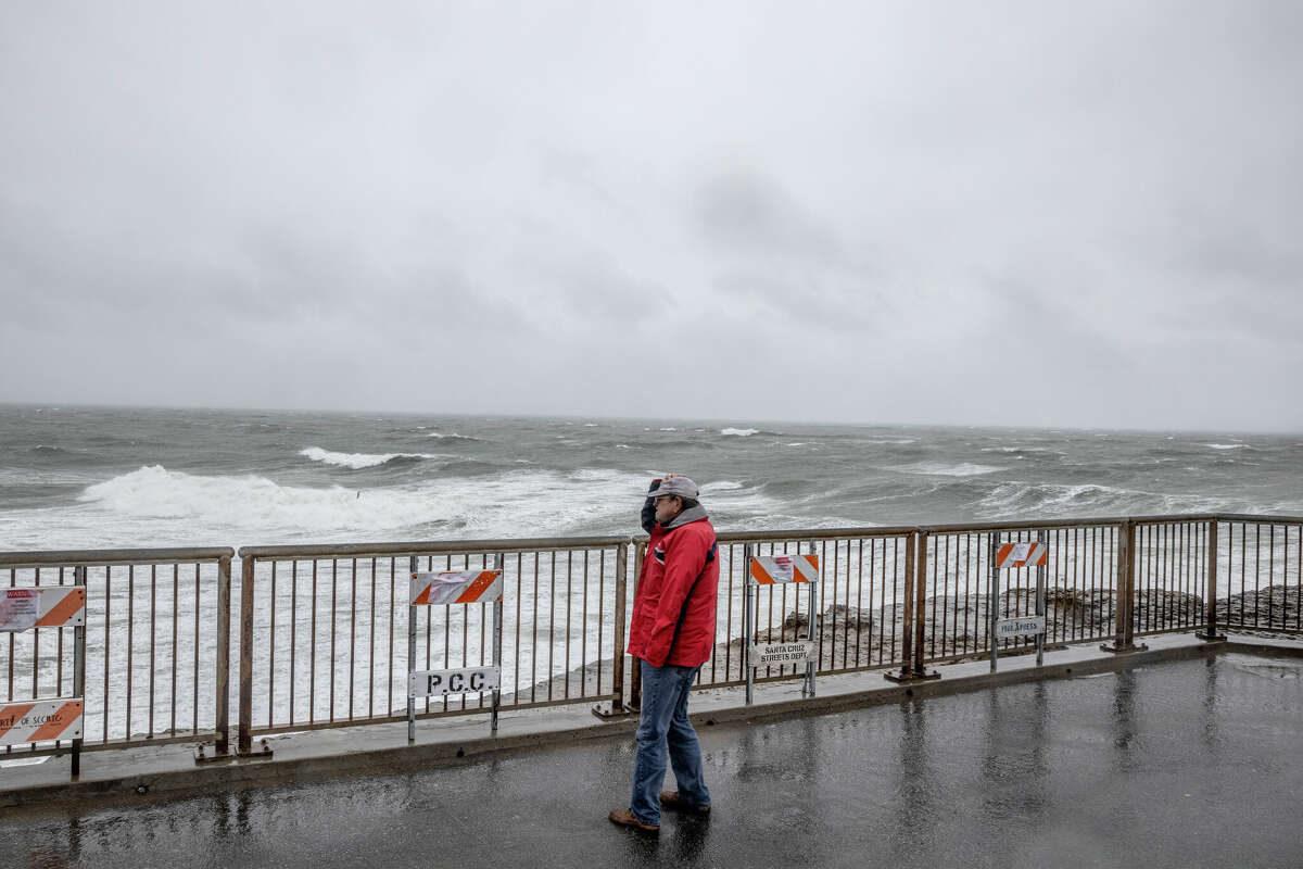周二，在圣克鲁斯最新的大气风暴事件中，一名男子在灯塔点观看海浪撞击海岸时拿着帽子。