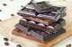 Broken chocolate bars of different kinds of chocolate are stacked on a wooden board, around a bit of coffee beans