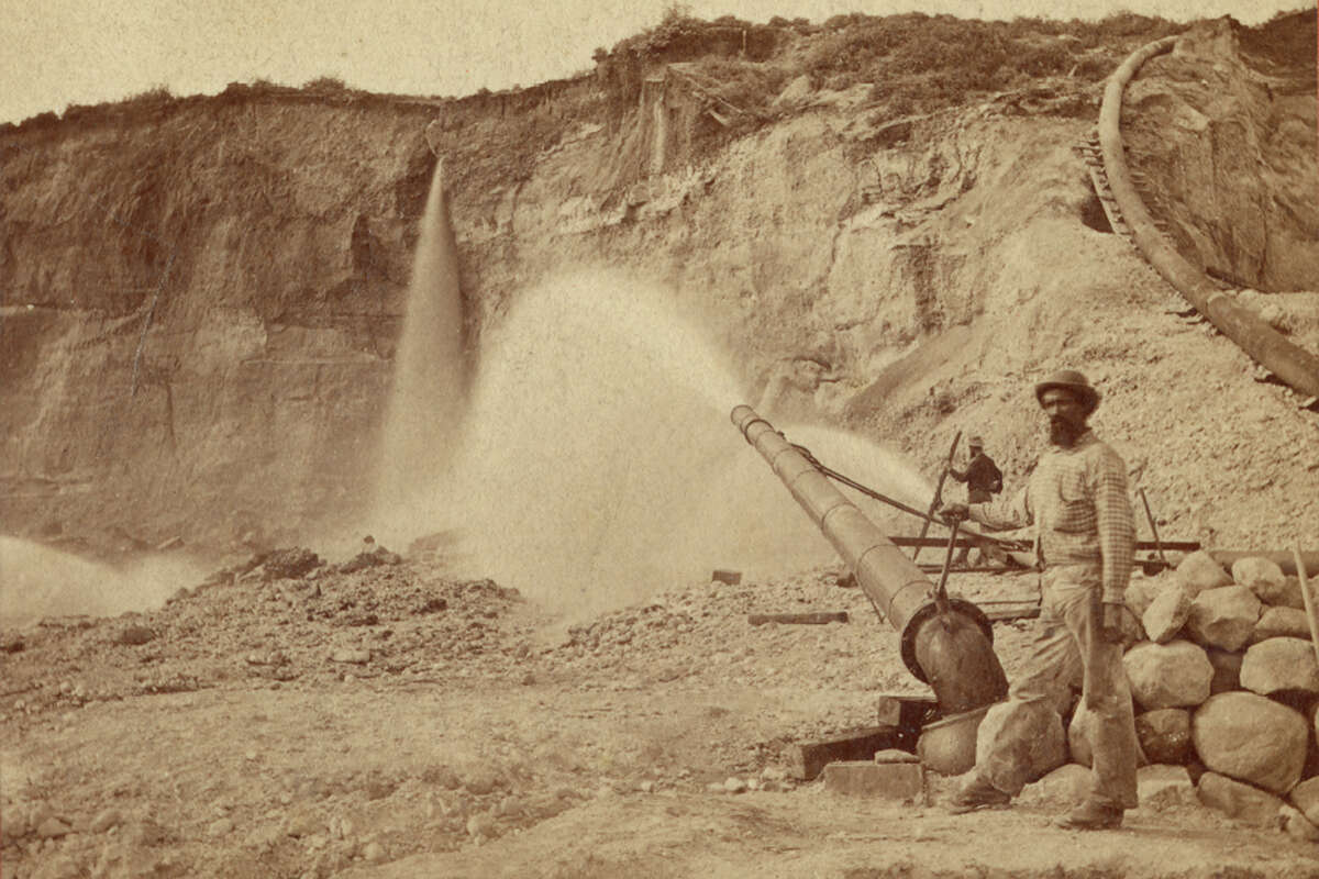 View of hydraulic mining at Malakoff Diggins by the North Bloomfield Gravel Mining Company in Nevada County; a miner is standing by a large monitor spraying water, 1874.