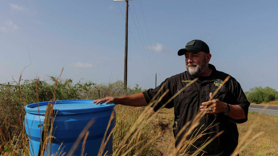 Jim Hogg County Sheriff's Investigator Ruben Garza inspects a water station for immigrants containing sealed jugs of fresh water along a fence line near a roadway in rural Jim Hogg County, Texas, Tuesday, July 25, 2023. The South Texas Human Rights Center maintains over 100 blue barrels consistently stocked with water across rural South Texas to serve as a life-saving measure for immigrants who have crossed into the United States to travel north in the sweltering heat.