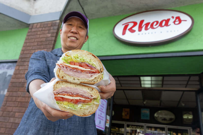 A man in a cap holds out the two halves of a deli sandwich under Rhea’s oval sign on its green storefront