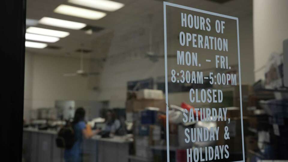 Post office hours are displayed in front of a line of people on Friday, Dec. 8, 2023, in Houston. Post offices are stopping service promptly at 5 pm no matter how many people are still in line.