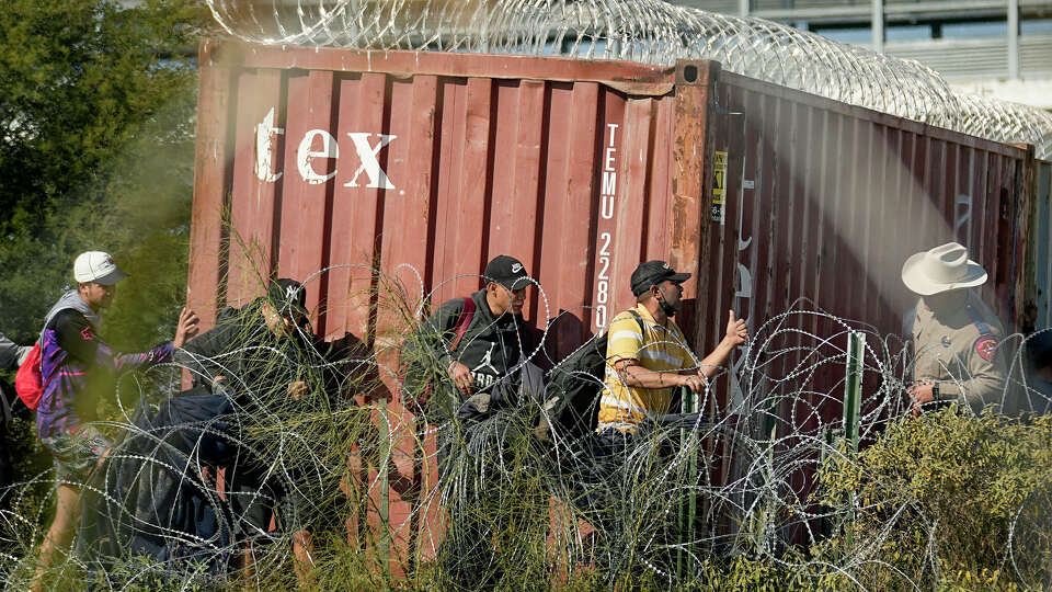 A Texas Department of Public Safety official, right, looks on as migrants walk near a rail car covered in Concertina wire at the Texas-Mexico border, Wednesday, Jan. 3, 2024, in Eagle Pass, Texas. U.S. House Speaker Mike Johnson is leading about 60 fellow Republicans in Congress on a visit to the Mexican border. Their trip comes as they are demanding hard-line immigration policies in exchange for backing President Joe Biden's emergency wartime funding request for Ukraine. (AP Photo/Eric Gay)