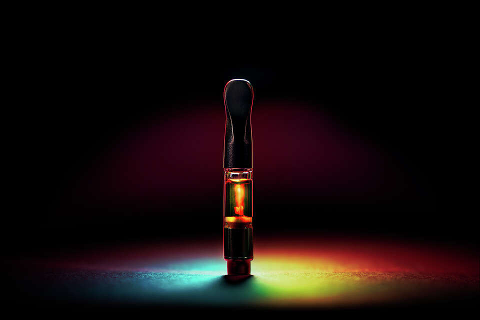 FILE: Marijuana vape pen cartridge with radiant rainbow lighting and dark background.