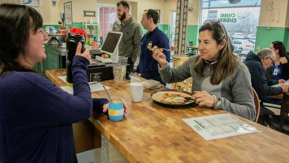 Teresa Panico lifts a fork and Daiana Goldstein does the same with a drink as Goldstein's husband Diego (center) chats with a customer in the background at new Milford restaurant Chimichurri on Wednesday, March 6, 2024.