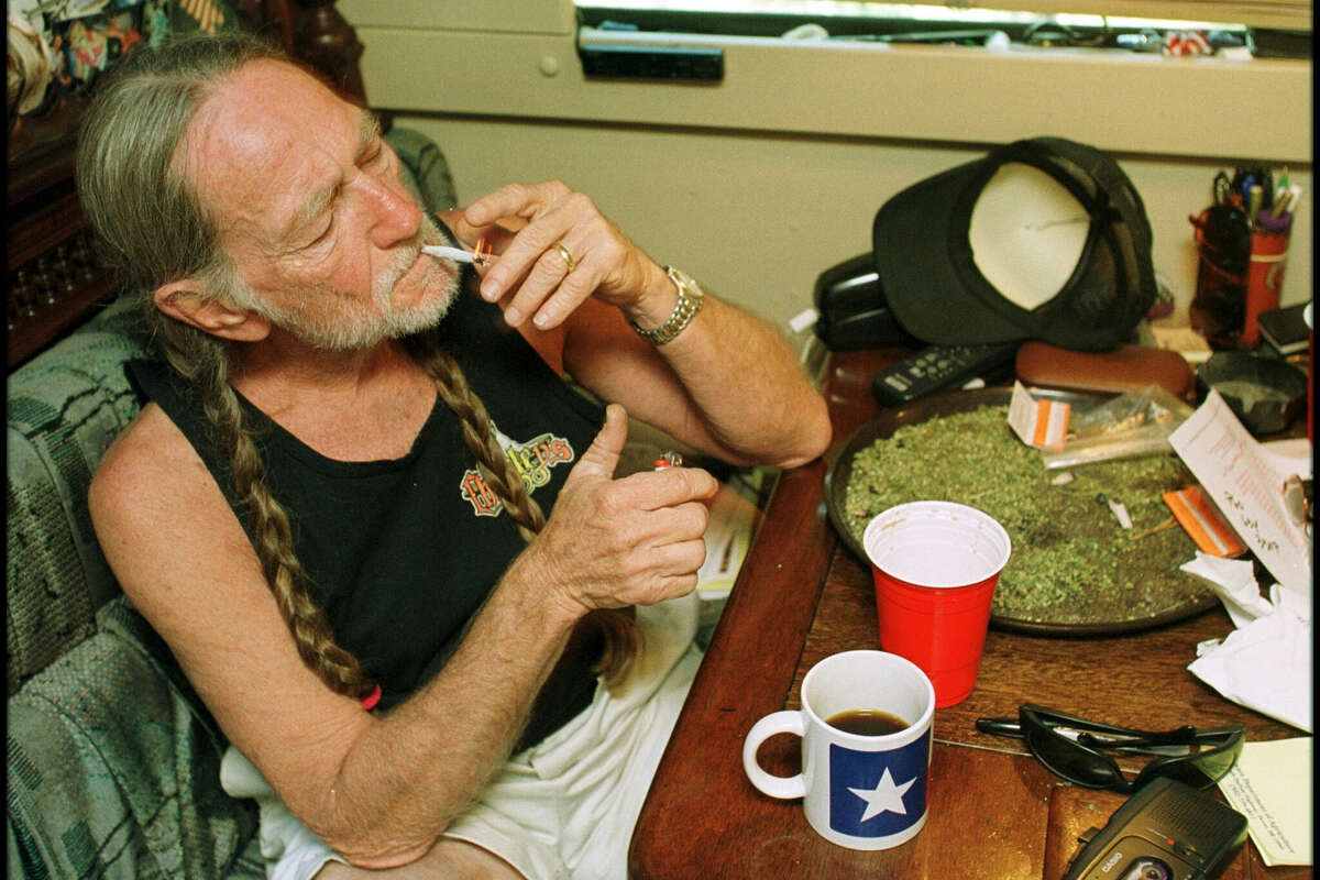 Willie Nelson takes a drag off a joint while relaxing at his home in Texas, 2000s. A large amount of marijuana is spread out on the table before him.