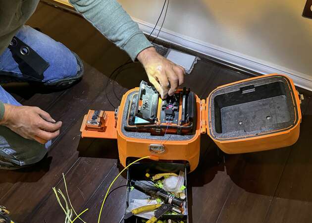 An AT&T fiber installer uses a device to splice two ends of fiber-optic cable. The device uses intense heat to melt the glass filaments, and a small screen shows the whole process. 
