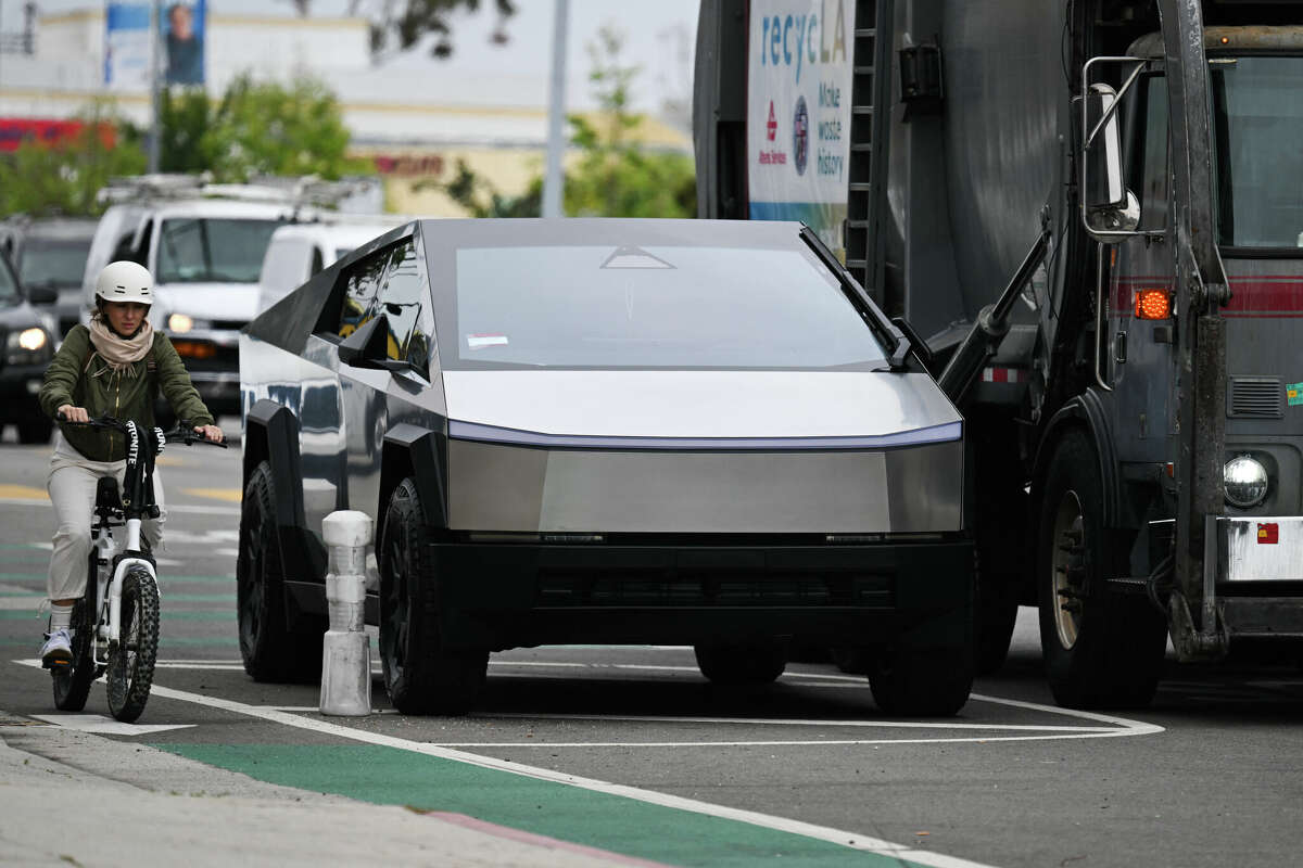 A cyclist rides past a Tesla Cybertruck parked next to a garbage truck in Los Angeles, California, on May 15, 2024. The electric vehicle's front wiper rests vertically on the side of the windshield.