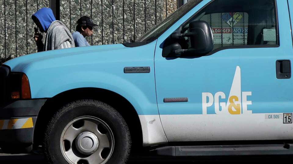 In this photo taken Feb. 18, 2020, people walk behind a Pacific Gas and Electric truck parked in San Francisco. The stock market turmoil triggered by the coronavirus pandemic is raising worries that Pacific Gas & Electric's $13.5 billion settlement with victims of catastrophic wildfires may be worth far less by the time the beleaguered company emerges from bankruptcy. A lawyer who represents more than 81,000 wildfire victims flagged the escalating concerns during a Wednesday, March 25, 2020, court hearing held by conference call. (AP Photo/Jeff Chiu)