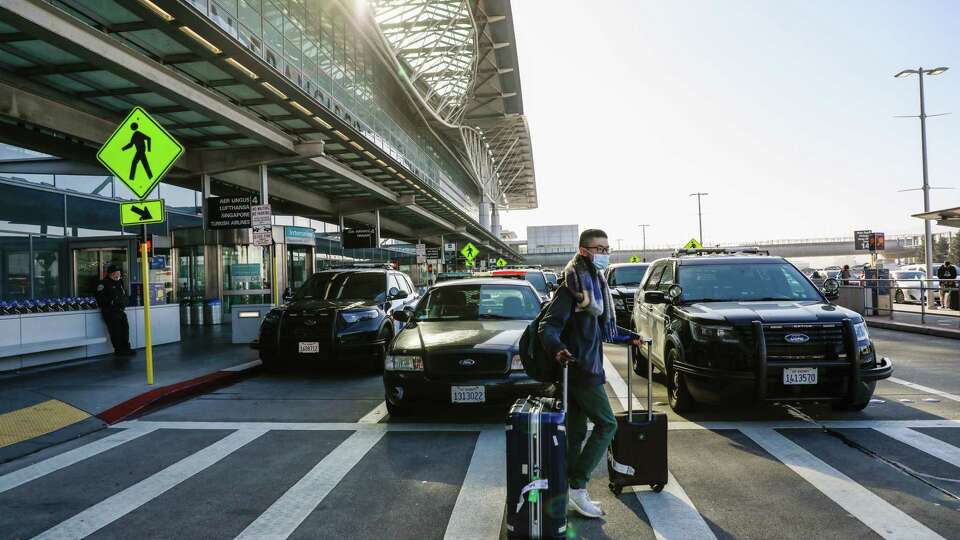 A traveler passes by police cars as they make their way out of the International Terminal at San Francisco International Airport (SFO) after an incident involving an armed individual in front of the BART station entrance in San Francisco, Calif., Thursday, Jan. 20, 2022. The suspect, allegedly armed with two guns, was fatally shot after officers were unable to de-escalate and non-lethal measures were ineffective, authorities said.