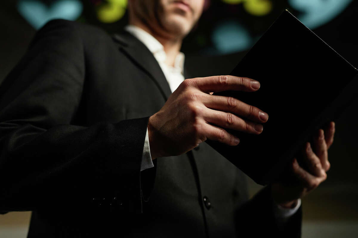 Close-up of mature man in black suit reading Bible standing in church (Stock image).