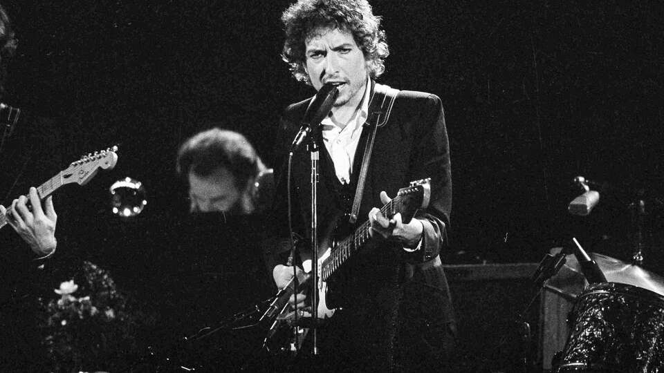 FILE - Musician Bob Dylan belts out a tune as members of The Band, Robbie Robertson, partially hidden left, and Garth Hudson, background center, accompany him, during a concert at the Forum in Los Angeles, Calif., Feb. 15, 1974. Columbia Records and Legacy Recordings announced Tuesday, July 9, 2024, plans to release a 27-disc CD box set of Dylan's memorable 1974 concert tour with The Band on Sept. 20. (AP Photo/Jeff Robbins, File)