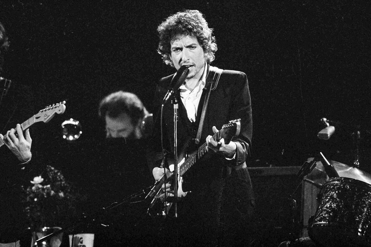 FILE - Musician Bob Dylan belts out a tune as members of The Band, Robbie Robertson, partially hidden left, and Garth Hudson, background center, accompany him, during a concert at the Forum in Los Angeles, Calif., Feb. 15, 1974. Columbia Records and Legacy Recordings announced Tuesday, July 9, 2024, plans to release a 27-disc CD box set of Dylan's memorable 1974 concert tour with The Band on Sept. 20. (AP Photo/Jeff Robbins, File)