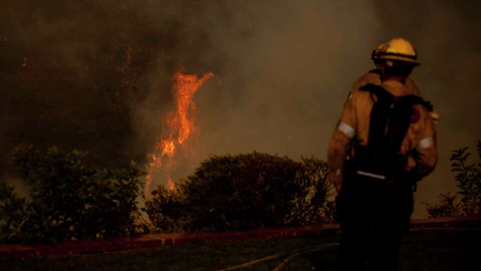 Firefighters watch as flames from the Palisades Fire are seen in the distance in the Mandeville Canyon neighborhood of Los Angeles,, Calif., Saturday, Jan. 11, 2025.