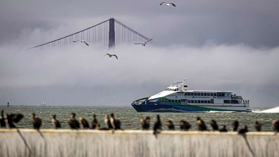 The San Francisco Bay Ferry passes by cormorants on a breakwater as clouds and fog shroud the Golden Gate Bridge in San Francisco Bay as seen from Treasure Island in San Francisco, on Wednesday, Sept. 18, 2024,