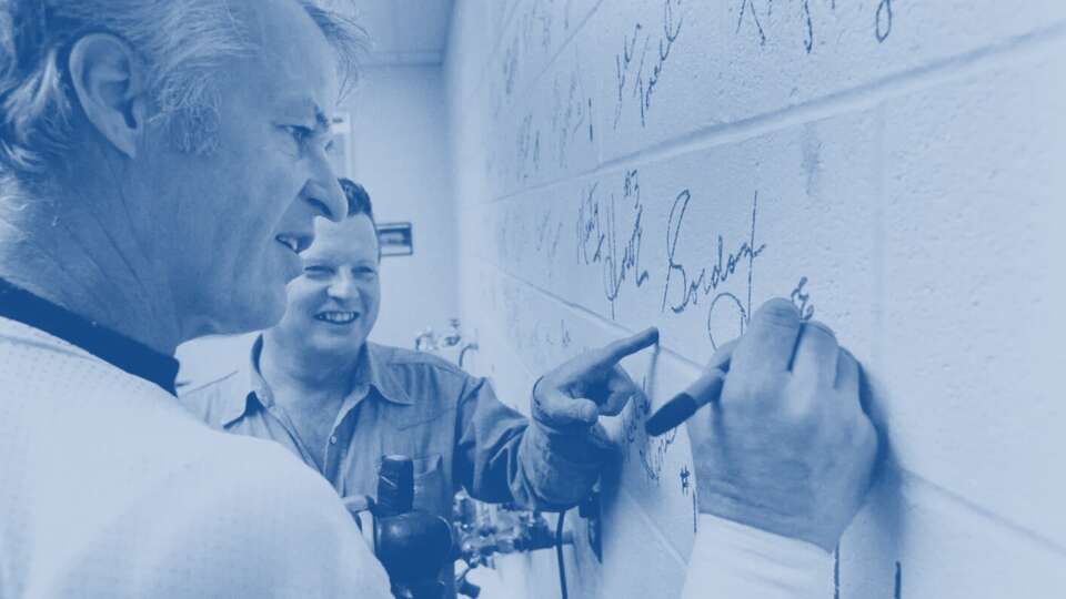 12/11/1975 - Gordie Howe adds the finishing touches to Bobby Brown's 'piece of the rock' wall Thursday, Dec. 11, as Brown, the Houston Aeros trainer, looks on. Each Aero signed a separate cinderblock in the training room wall, signifying the team's role in helping make Houston's new arena, The Summit, possible.