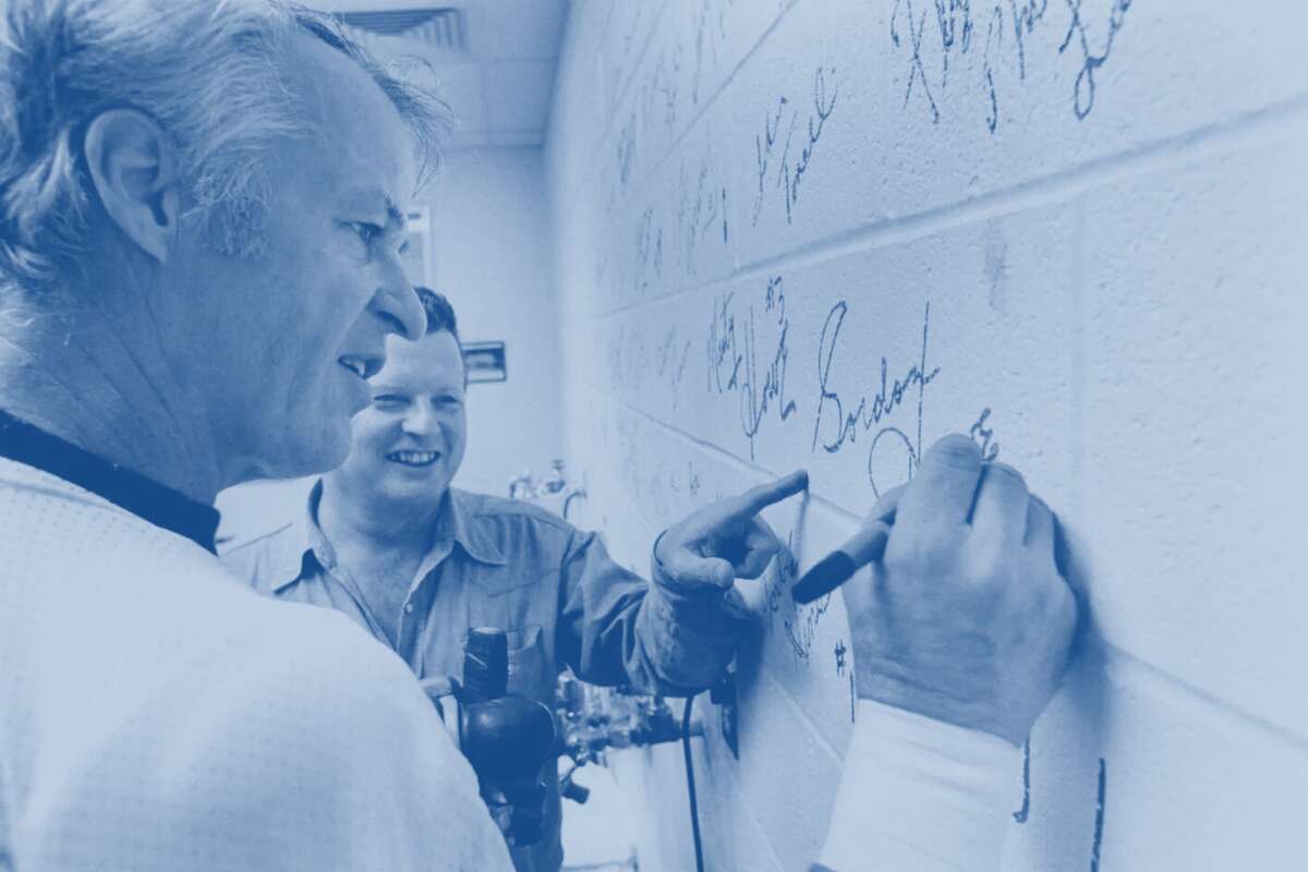 12/11/1975 - Gordie Howe adds the finishing touches to Bobby Brown's 'piece of the rock' wall Thursday, Dec. 11, as Brown, the Houston Aeros trainer, looks on. Each Aero signed a separate cinderblock in the training room wall, signifying the team's role in helping make Houston's new arena, The Summit, possible.