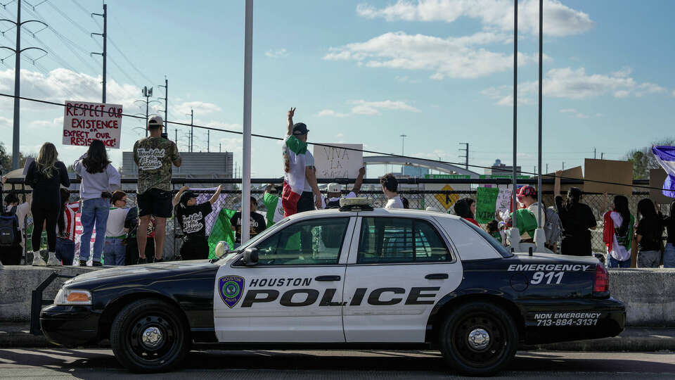 A Houston Police car is seen parked in front of people holding signs and waving flags in protest of the ongoing ICE raids on Sunday, Feb. 2, 2025 in Houston.