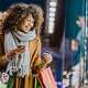 Smiling woman wearing winter coat and scarf holding shopping bags and using her cell phone as she shops for gifts while outside store front window.