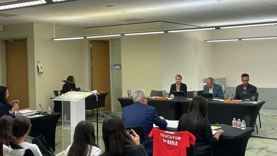 Jamie Russo, a 38-year teaching veteran seated on the far right, begins her hearing on Nov. 20, 2025 before Houston ISD's appointed Board of Managers. Over the back of her chair is a t-shirt that reads 'HISD educator in exile.' To Russo's immediate left is attorney Chris Tritico.
