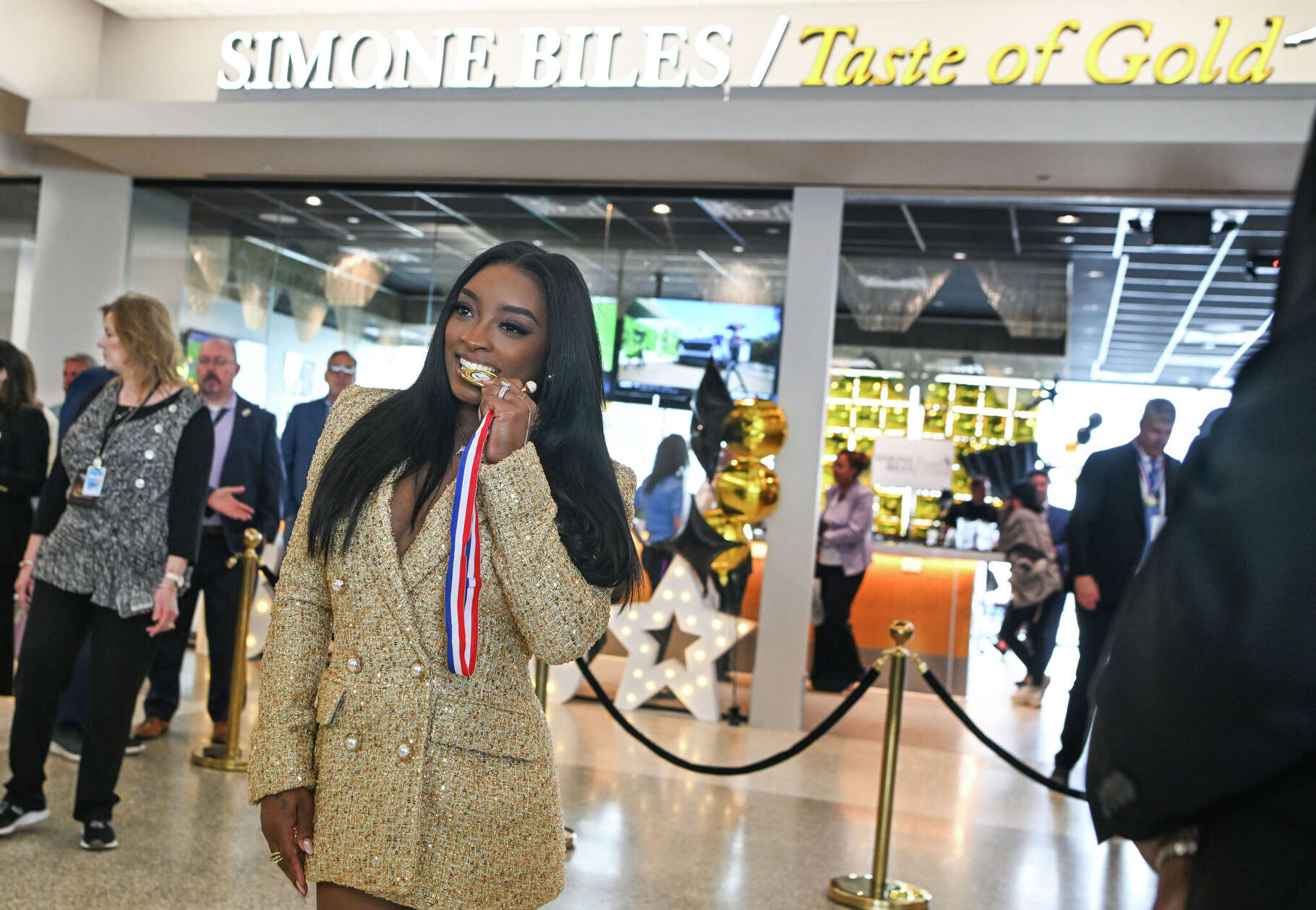 Simone Biles poses bites a gold medal while celebrating the opening of her 'Taste of Gold' restaurant at George Bush Intercontinental Airport in Houston on Tuesday, March 24, 2026.