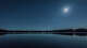 A bright moon shines over a calm lake at night, with stars visible above and reflections shimmering on the water.