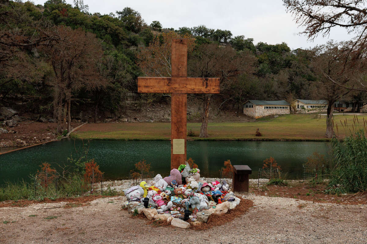 A memorial cross is displayed beside Texas 39, with Camp Mystic cabins visible across the Guadalupe River in Hunt, Texas, on Wednesday, Dec. 3, 2025.