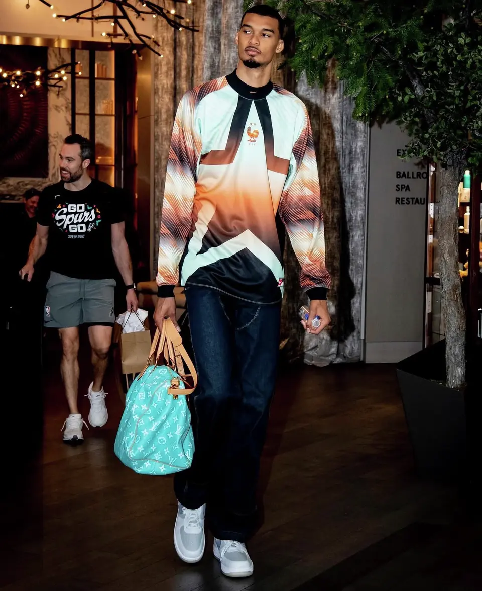 Victor Wembanyama arrives in a Nike France “Hollywood” goalkeeper jersey paired with loose denim and a light blue Louis Vuitton bag, blending sport and luxury in a signature tunnel look.