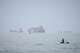 A man sits on a surfboard as a container ship and other commercial vessels appear anchored in the Strait of Hormuz off Bandar Abbas, Iran, Monday, July 27, 2026. (Razieh Poudat/ISNA via AP)