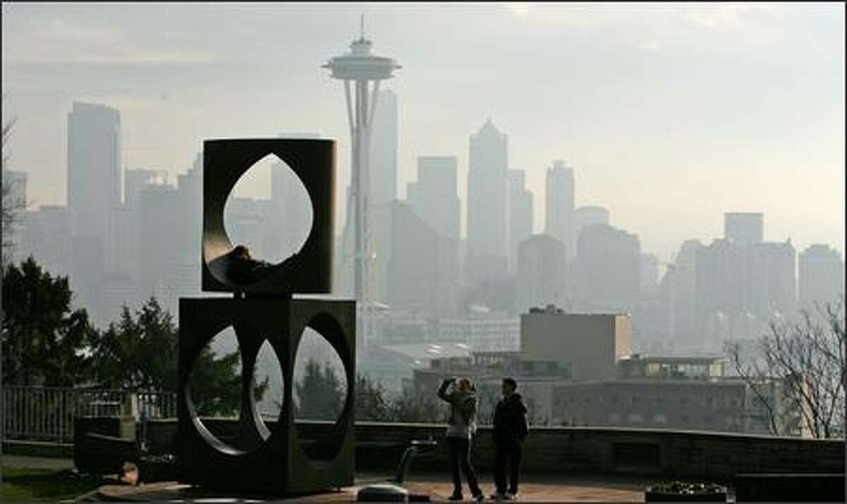 Haze covers Seattle as Michelle Chavez, center, photographs Jonathan Hopper in the sculpture at Kerry Park. Aaron Lum, right, watches. Many residents who still don't have power are burning wood to keep warm, which is causing regional air quality to worsen.