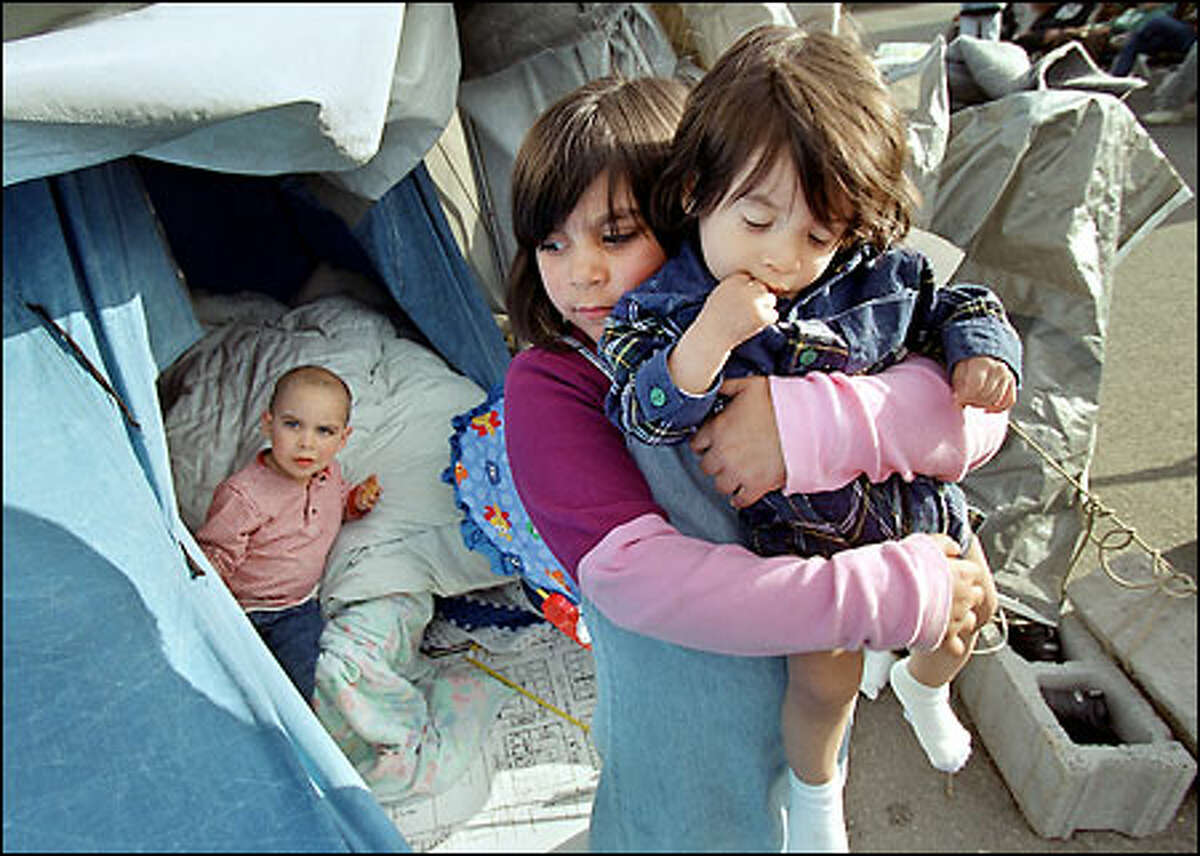 Kennedy Zuniga, 6, lifts Kailebh, 11 months, as her brother Skylar, 3, peers from their tent. They are living at Tent City with their mother, Rachel Zuniga.