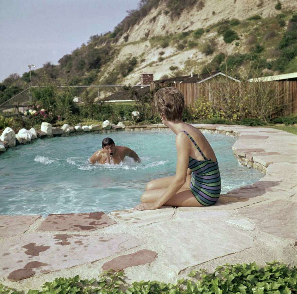 While wife Margaret sits on the edge of the pool between swims, actor Clint Eastwood thrashes about, enjoying the refreshing dip. In back is their home, Oct. 25, 1962 in Hollywood Hills, Calif.