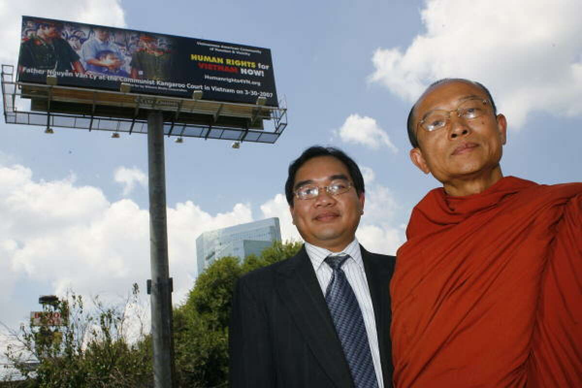Van Huynh and the Rev. Huyen Viet, a Buddhist monk, stand before a billboard depicting Father Nguyen Van Ly being prevented from speaking in a Communist peoples' court.