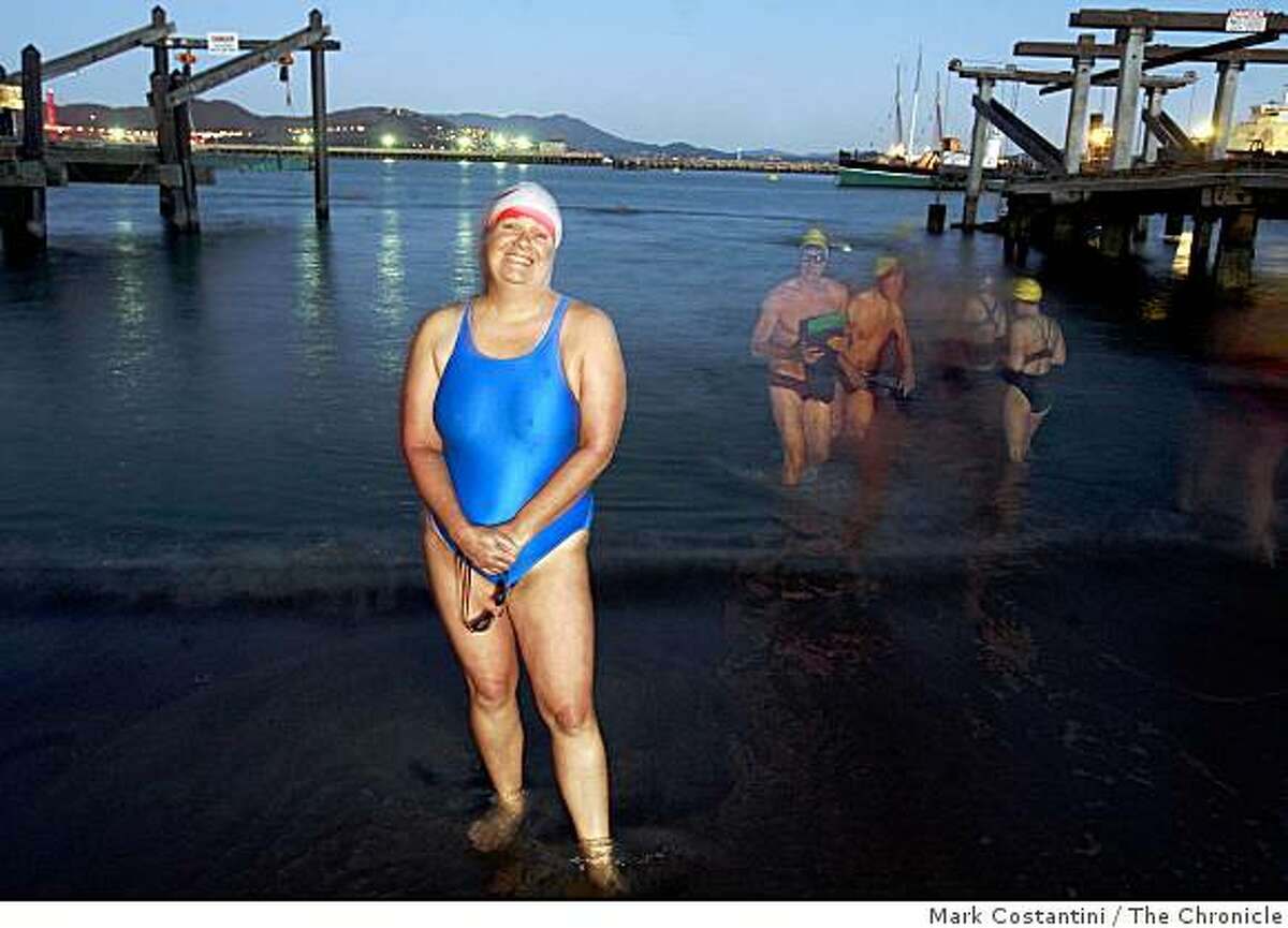 In the dark, before sunrise, when she practices, Suzie Dods, 48, an open water swimmer stands at the water's edge at the Dolphin Club in San Francisco, Calif. Behind her are other swimmers leaving the water after they are finished practicing.