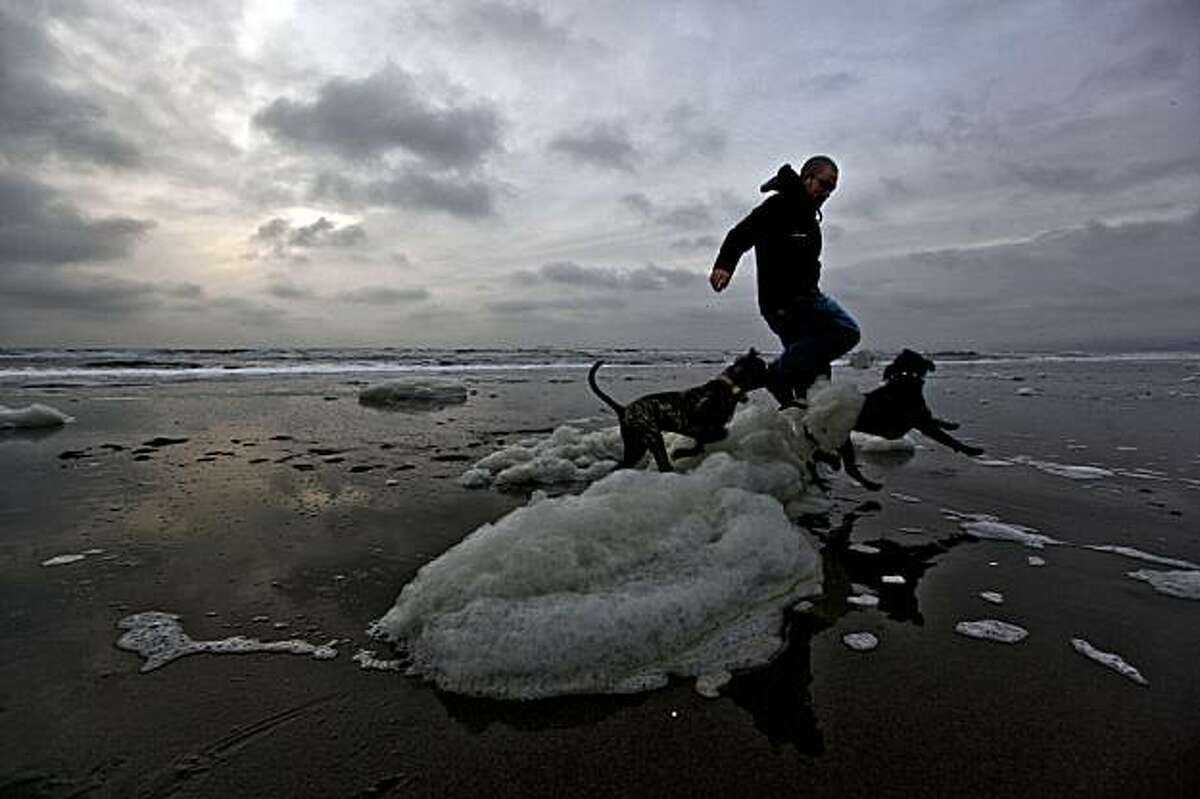 Alan Rodriguez, of San Francisco, hurdles a little beach foam with his dogs, "Bruiser" (left) and "Oynx" along ocean beach towards the end of the day as thick cloud cover rolls onshore on Friday December 4, 2009.