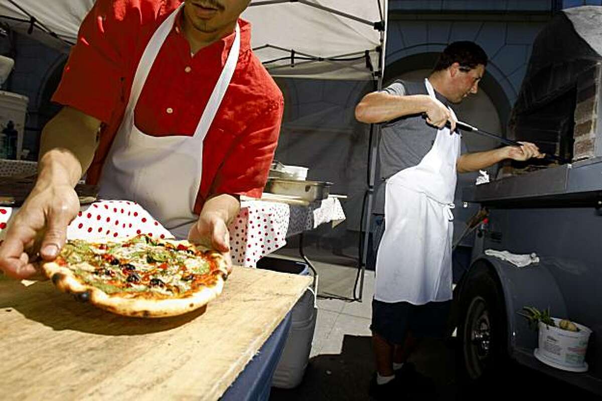 Dung Tran (left) cashier, prepares a pizza for a customer as lead cook, Aaron Lerch (right), monitors pizzas in the oven at the Pizza Politano booth at the Ferry Building Farmers' Market in San Francisco, Calif. on Thursday, August 13, 2009.