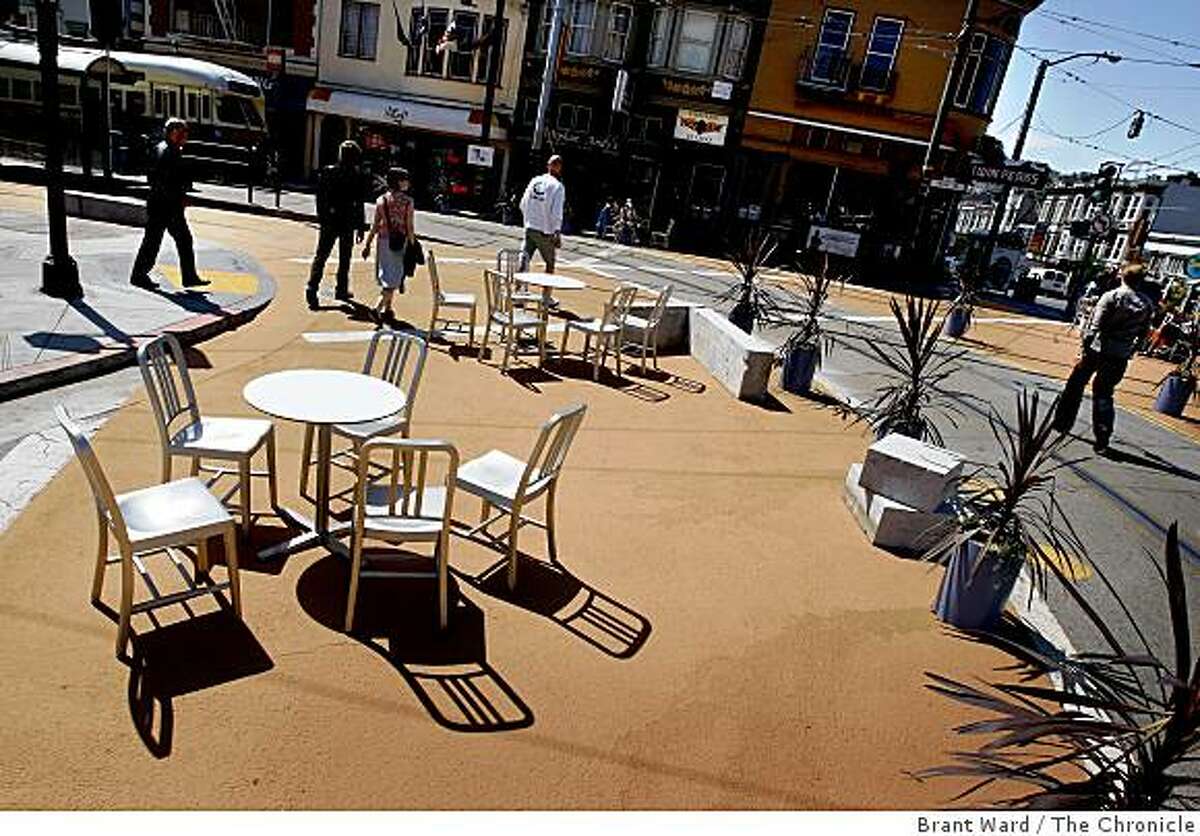 Tables and chairs cast shadows in the morning light as pedestrians make their way through the plaza. A temporary public plaza at the end of the streetcar line at 17th Street and Castro Streets in San Francisco, CA creates a place where people can linger and watch the world go by.