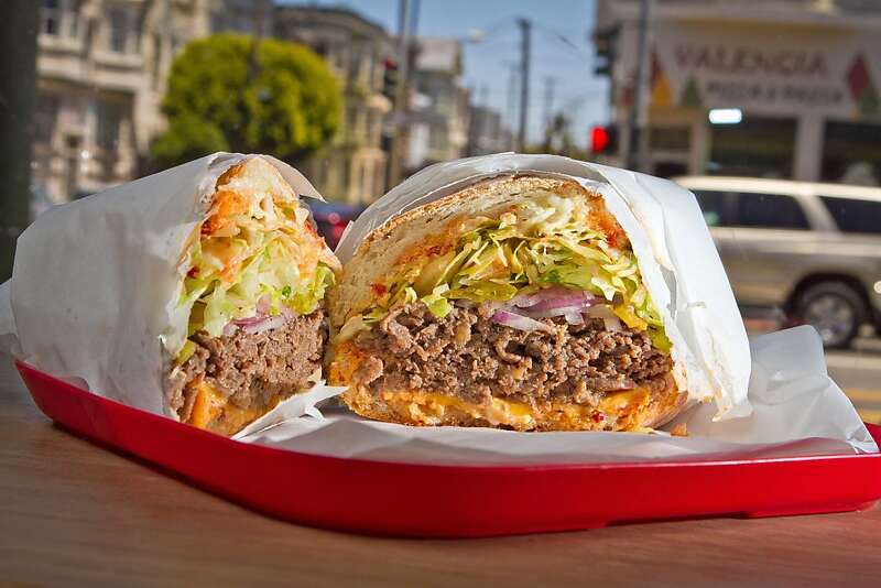 A steak sandwich cut in half in a red tray, with shredded lettuce, red onion and an orange sauce, Valencia Street behind it
