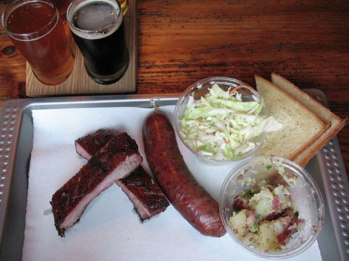 A two-meat lunch plate at The Granary has ribs and sausage, with sides of cole slaw and German potato salad. Buttermilk bread is available separately. In the background is a flight of four house brews.