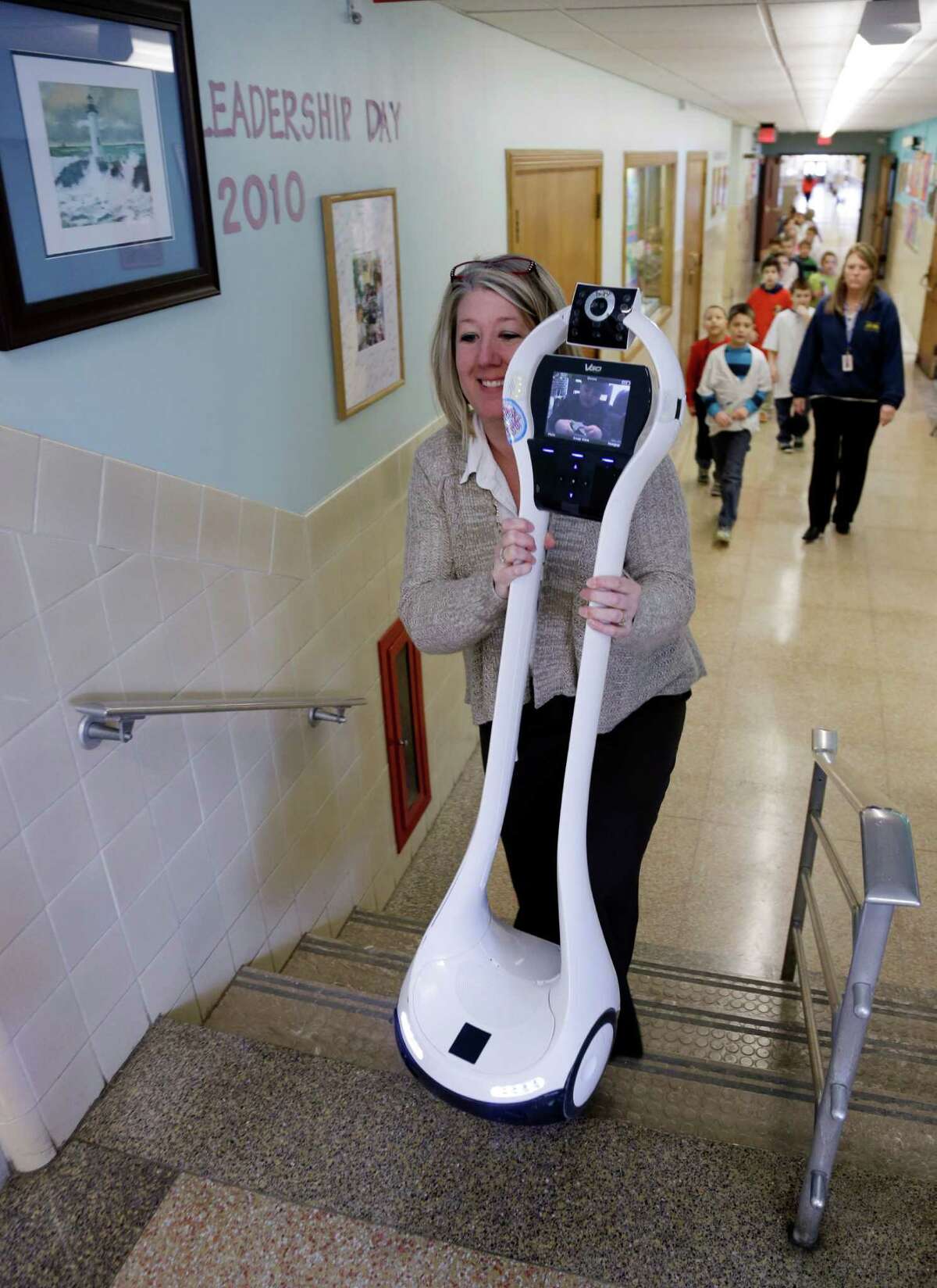 In this Thursday, Jan. 24, 2013 photo, Winchester Elementary School teacher Dawn Voelker carries a robot up the stairs that is being operated by Devon Carrow while he is attending school at in West Seneca N.Y. Carrow's life-threatening allergies don't allow him to go to school. But the 4-foot-tall robot with a wireless video hookup gives him the school experience remotely, allowing him to participate in class, stroll through the hallways, hang out at recess and even take to the auditorium stage when there's a show. (AP Photo/David Duprey)