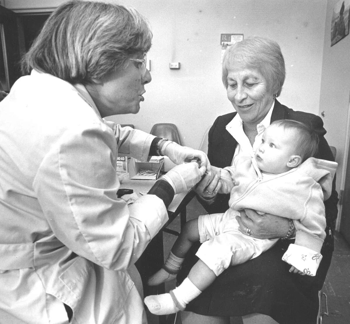 Lee Stoogenke, a lab technician for the Stamford Health Department, does a blood test on 9-month-old Sheila Coperine, whose mother was a teacher at K.T. Murphy School, which was closed due to high lead levels. Holding Sheila is Springdale School nurse Estelle Skigen.