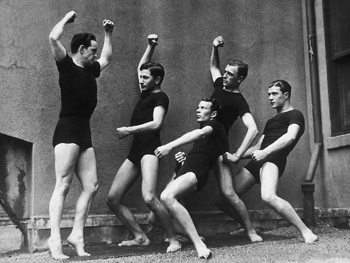 Full-length image of five men, dressed in matching black T-shirts and shorts, contorting their bodies while exercising, possibly on a rooftop, 1910s. The leader holds his stomach in and flexes his muscles while the others watch.