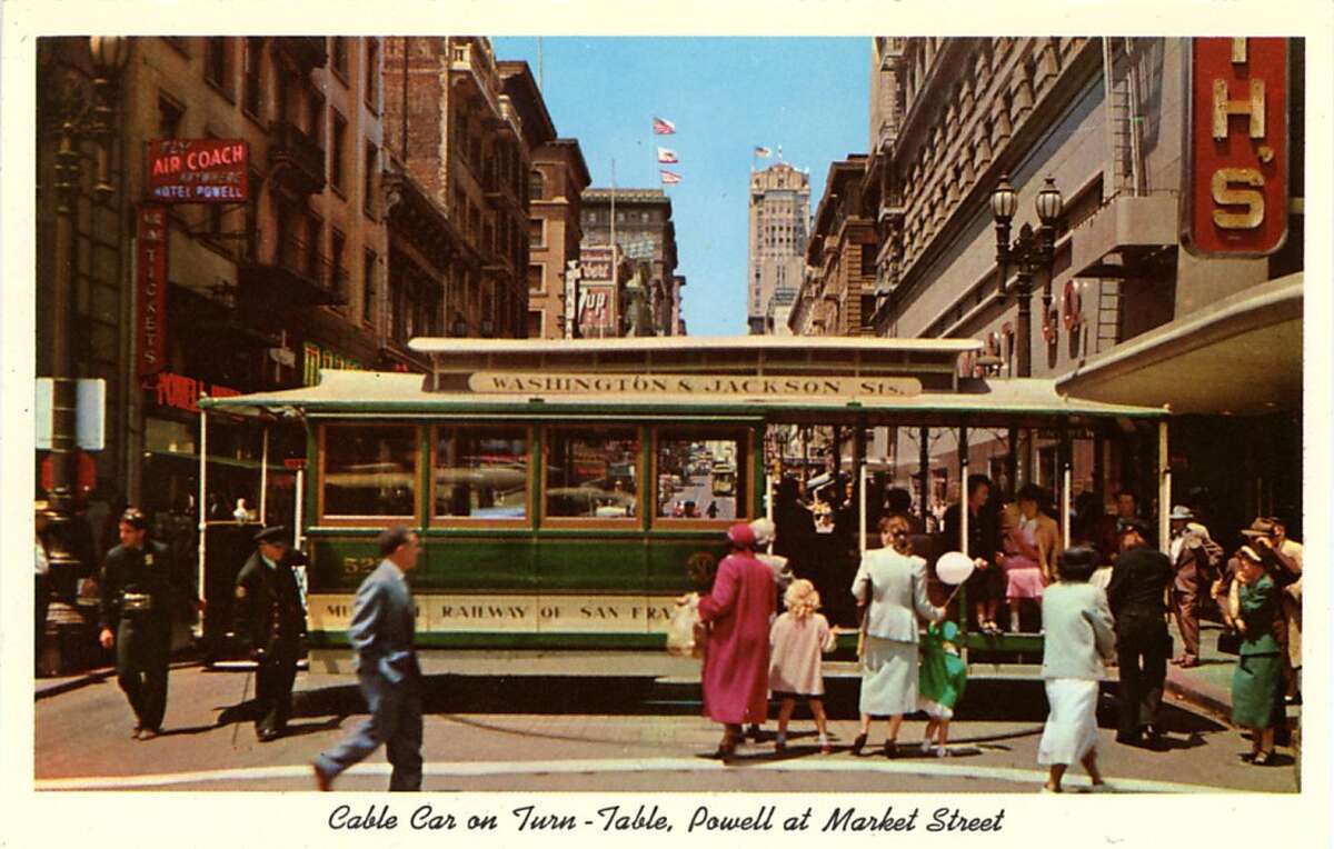 Vintage postcard showing a cable car on the turntable in downtown San Francisco. People wait to embark the car and the cityscape is visible in the background.