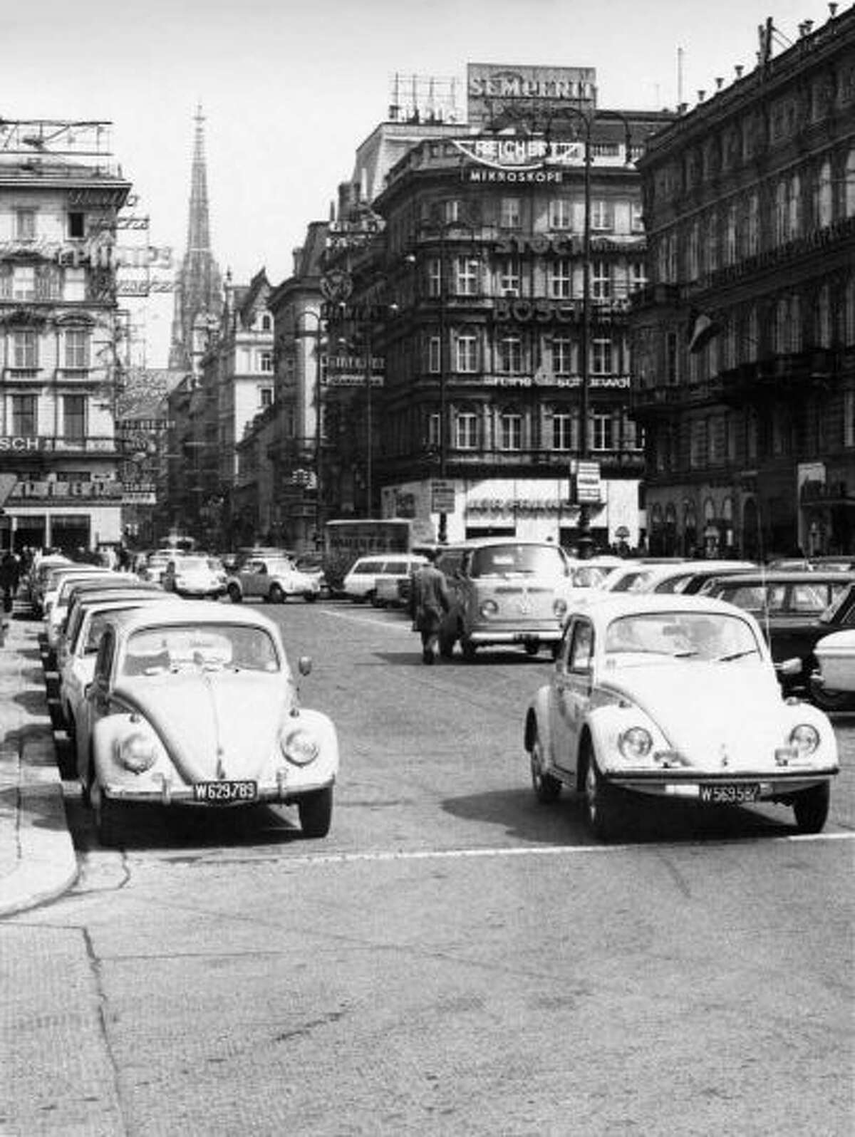 A Volkswagen Type 2 van putters along in the background behind Volkswagen Type 1 cars (aka Beetles) in Vienna, Austria, 1965.