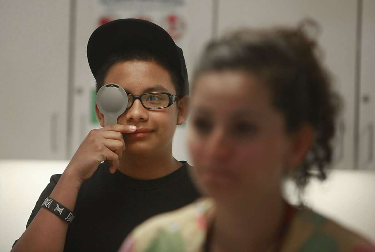Carlos Aguilar (left), 14, prepares to take an eye exam from Eneida Vera (right), medical assistant, during a physical at the Havenscourt Health Clinic on Wednesday, July 10, 2013 in Oakland, Calif.