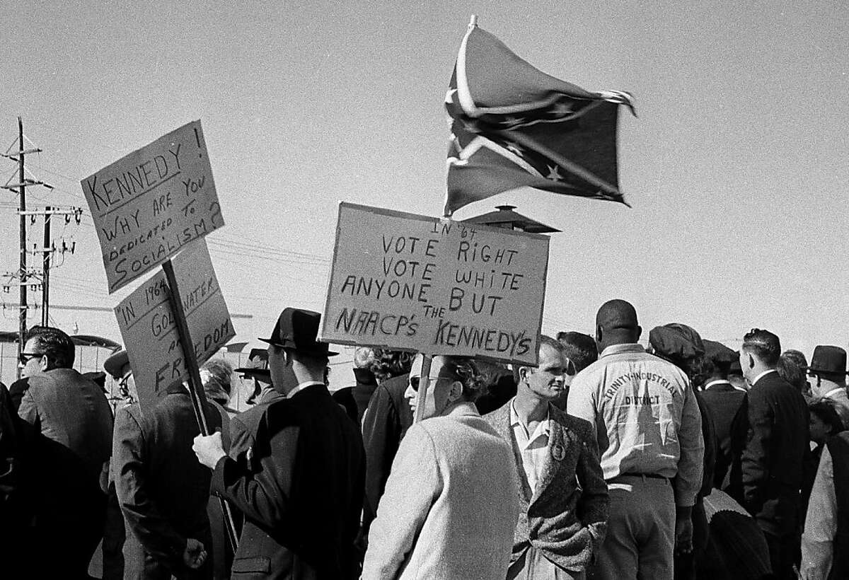 ADVANCE FOR USE SUNDAY, JULY 14, 2013 AND THEREAFTER - FILE - In this Nov. 22, 1963 file photo, people, including right-wing protesters carrying a Confederate flag and anti-Kennedy placards, await the arrival of President John F. Kennedy at Love Field in Dallas. The 50th anniversary of Kennedy's assassination throws a new spotlight on the deep hostility toward Kennedy that some Dallasites voiced before the assassination. (AP Photo/File)
