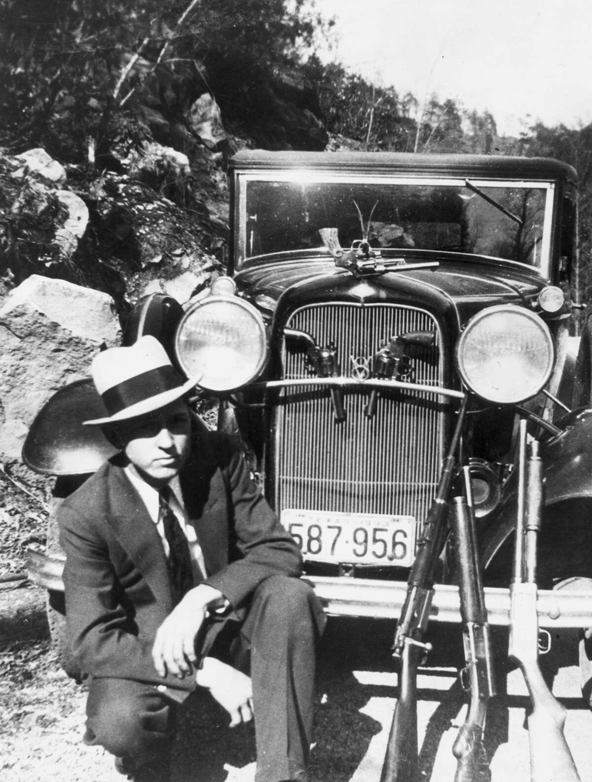 Clyde Barrow crouches in front of a car with a handgun hanging from the hood ornament, two handguns in the front grill and three rifles laid against the front fender, possibly in Joplin, Mo., in April 1933.
