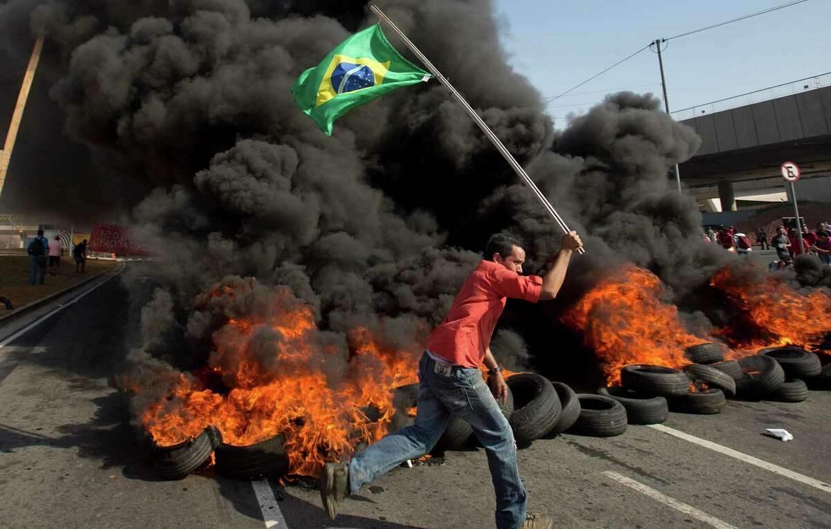 In this May 15, 2014 photo, a member of the Homeless Workers Movement carries a Brazilian flag past burning tires during a protest against the money spent on the World Cup near Itaquerao stadium which will host the international soccer tournament's first match in Sao Paulo, Brazil. Brazilians are angry at the billions spent to host the World Cup, much of it on 12 ornate football stadiums, one-third of which critics say will see little use after the big event. (AP Photo/Andre Penner,File)